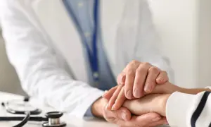 Lekarz trzyma dłoń pacjenta, oferując wsparcie w obliczu trudnych objawów raka. Stetoskop leży na stole.