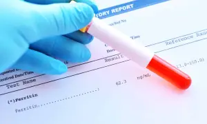 Ręka w niebieskiej rękawiczce trzyma probówkę z krwią nad raportem laboratoryjnym, gdzie widnieje wynik badania ferytyny.