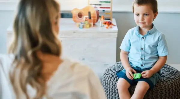 Chłopiec w niebieskiej koszuli siedzi na pufie, rozmawiając z psychologiem. W tle zabawki.