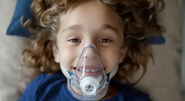 Uśmiechnięte dziecko z kręconymi włosami w masce inhalacyjnej.