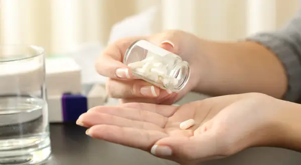 Kobieta wysypuje kapsułki z buteleczki na dłoń. Czy paracetamol jest przeciwzapalny? Obok stoi szklanka wody i pudełka leków.