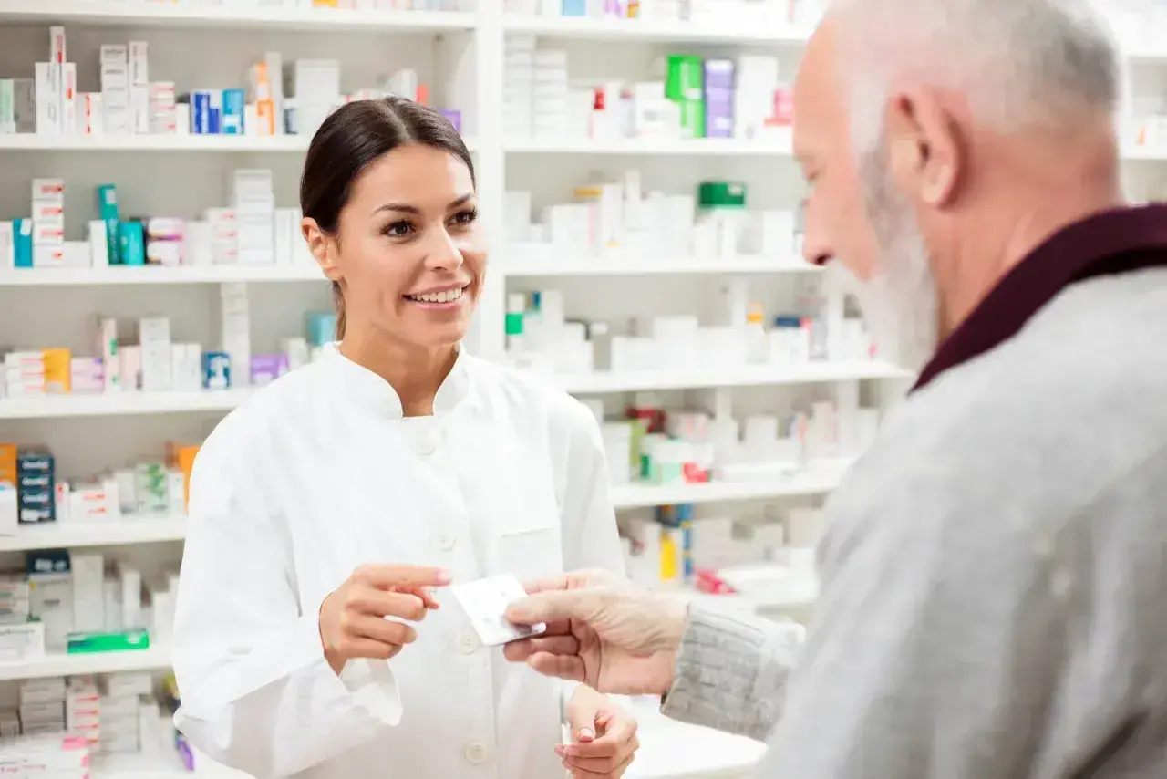 Farmaceutka podaje starszemu panu leki. W tle widać półki pełne opakowań, być może z eliquis.