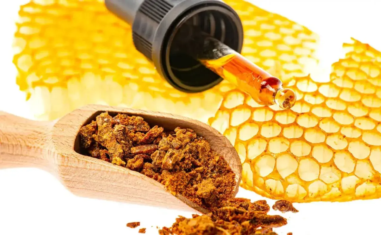 Propolis co to? Złota kropla z pipety na plastrze miodu, obok drewnianej łyżki z pyłkiem.