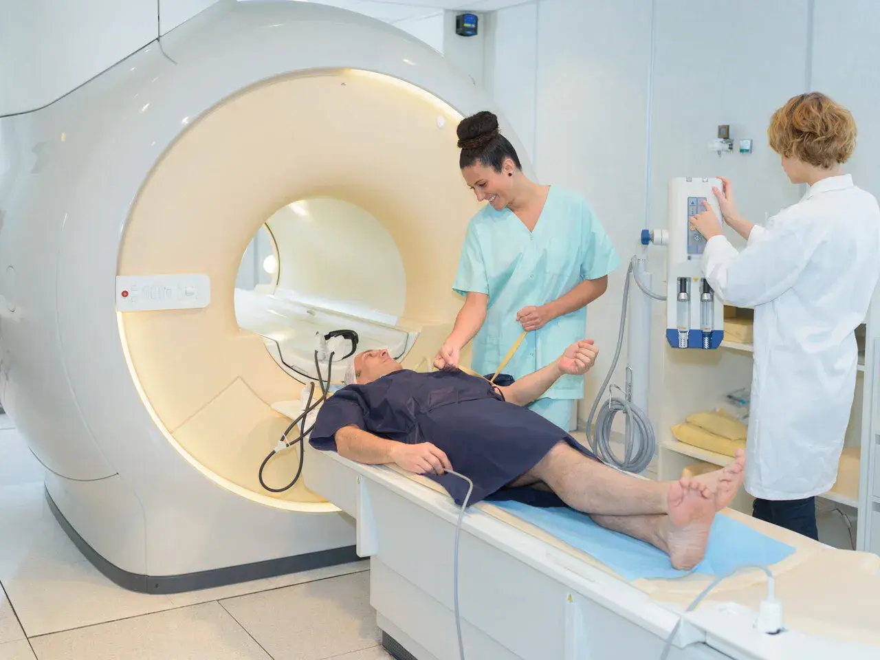 Pacjent leży na łóżku przed aparatem MRI. Dwie pielęgniarki przygotowują go do badania. To właśnie mri co to za badanie.