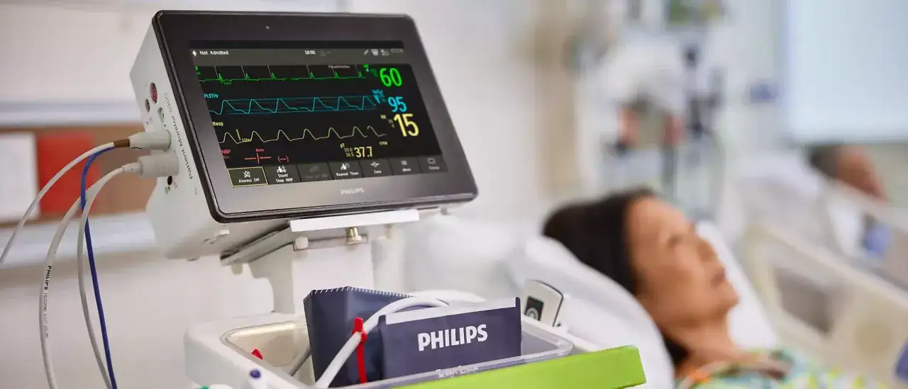 Monitor parametrów życiowych pacjenta, pokazujący EKG, oddech i ciśnienie. W tle pacjentka, być może po podaniu midazolamu.
