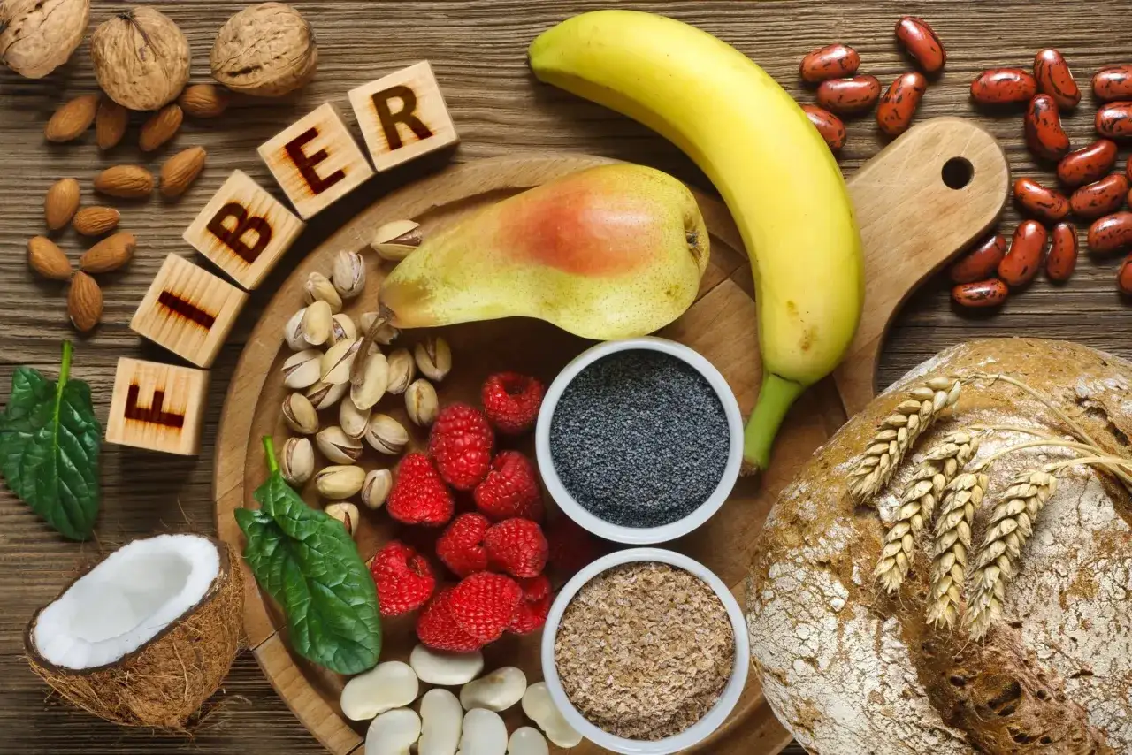 Drewniany stół pełen produktów bogatych w błonnik: orzechy, migdały, pistacje, maliny, banan, gruszka, fasola, mak, płatki owsiane, chleb i kokos. Zastanawiasz się, ile błonnika dziennie?
