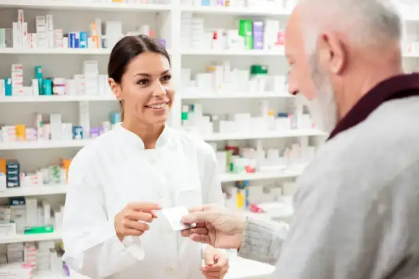 Farmaceutka podaje starszemu panu leki. W tle widać półki pełne opakowań, być może z eliquis.