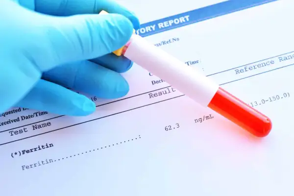 Ręka w niebieskiej rękawiczce trzyma probówkę z krwią nad raportem laboratoryjnym, gdzie widnieje wynik badania ferytyny.